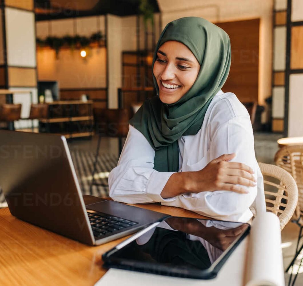 Muslim university student smiling happily while having a video conference with her tutor in a cafe. Cheerful woman with a hijab attending an online class in a coffee shop.